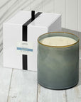 A large gray hand-blown glass vessel candle with three wicks, the Lafco NY Beach House Candle, sits on a white wooden surface beside its gift box tied with a black ribbon. Soft shadows fall across the scene.