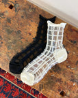 Two pairs of Unified Sheer French Window ankle socks, one black and one white, are displayed side by side on a rustic wooden surface near a patterned rug.