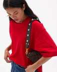 A woman in a loose red top and blue jeans looks down, carrying the Clare Vivier Shoulder Strap Naturelle W/ Studs—a small brown leopard-print bag with a black leather strap accented by shiny brass studs—against a plain white background.