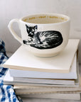The Sir/Madam Grand Cup, white with a black cat illustration and "CAT," "GATTO," and "CHAT" around the rim, rests atop books beside a blue and white checkered cloth.