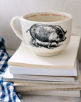 The Sir/Madam Grand Cup, a large white mug with a black pig illustration, rests on stacked books beside a blue and white checkered cloth. The rim features the animal's name in four languages for colorful repetition.