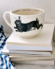 The Grand Cup by Sir/Madam, featuring a black rabbit illustration and "rabbit" in several languages around the rim, sits atop white books next to a blue and white checkered cloth.