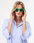 A woman wears bright green cat-eye sunglasses, a light blue button-up shirt, and the Clare Vivier Stone Slice Necklace as she poses adjusting her sunglasses against a plain white background.