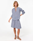 A woman with blond hair, a pearl necklace, and brown flat sandals wears the Clare Vivier Zoe Skirt—blue and white linen gingham with a smocked waist and pockets—standing against a plain white background.
