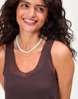 A woman with long, wavy dark hair smiles in a sleeveless brown top and layered necklaces, including Clare Vivier’s Stone Slice Necklace, against a light background.