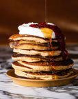 A stack of Wild Rye Almond Flour Pancake Mix pancakes on a golden plate, topped with whipped cream and red berry sauce, with syrup being poured over them, sits on marble against a warm brown background.