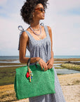 A woman in a black-and-white gingham dress and sunglasses stands on a beach, holding a large green woven bag adorned with the Clare Vivier Stone Slice Necklace. The ocean and sky are visible in the background.