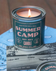 A G&W Soy Candle by Faire from the Happy Place Collection rests on a blue bandana and vintage camp photo. Its retro label and scent evoke nostalgic, carefree summer memories.