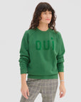 A woman with wavy brown hair wears a green Clare Vivier Sweatshirt / Flocked OUI made from 100% cotton and plaid pants, standing against a plain white background, looking slightly to the side and smiling softly.