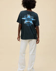 A person with short curly hair faces away, wearing the DayDreamer Backstreet Boys Into The Millennium oversized tee featuring "Into the Millennium" with a globe and star on the back, light pants, and black shoes—a tribute to classic band merch.