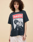 A young woman with curly hair wears the DayDreamer Oasis Merch Tee—an oversized black shirt with a photo of two men and the band's name in bold red letters—paired with light-colored pants, against a beige background.