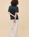 A person with curly hair faces away, wearing the oversized Oasis Merch Tee by DayDreamer, paired with white pants and black shoes, one hand behind their back against a neutral beige background.