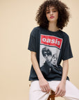 A young person with curly hair wears the DayDreamer Oasis Merch Tee—an oversized cotton shirt with a red logo and black-and-white image of two men—paired with light pants while sitting on a wooden stool against a beige background.