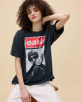 A person with curly hair wears the oversized DayDreamer Oasis Merch Tee and light pants while sitting on a wooden stool against a beige background, resting one hand behind their head.