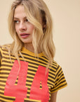 A woman with wavy blonde hair wears the DayDreamer 44 Striped Vintage Tee in yellow and brown with a bold red "44" on the front, standing against a plain beige background and looking to her left.