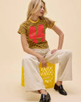 A woman with long blonde hair sits on a yellow crate, wearing the DayDreamer 44 Striped Vintage Tee, cream wide-leg pants, and black loafers. She looks to her left against a beige background.