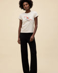 A woman with short curly hair wears DayDreamer's Ski Ya Later Tee and black wide-leg pants, standing against a plain beige background.
