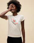 A smiling woman with curly hair stands against a beige background in the DayDreamer Ski Ya Later Tee—a women’s cotton t-shirt featuring a playful graphic and the words “SKI YA LATER.” She covers part of her face with one hand, looking relaxed and happy.