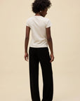 A person with short curly hair stands facing away, wearing the DayDreamer Ski Ya Later Tee, loose black pants, and black shoes against a plain beige background.