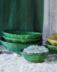 A set of hand-shaped ceramic bowls from Blue Ocean Traders, featuring mostly green and one yellow Tamegroute Bowl, are stacked on a white cloth. The smallest bowl in front displays round, translucent white beads as decoration.