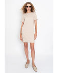 A woman with curly hair wears Kule's The Tee Dress—a beige and white striped, 100% cotton short sleeve dress—paired with sunglasses and light-colored moccasin shoes against a white background.