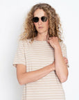 A person with curly blonde hair wears sunglasses and the Kule The Tee Dress—a beige, short-sleeved, white-striped 100% cotton dress—standing against a plain white background with arms crossed and one hand on their shoulder.