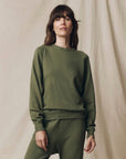 A woman with brown hair, wearing The College Sweatshirt S25 by The Great Inc. and matching pants in olive green, stands smiling gently at the camera before a beige fabric backdrop.