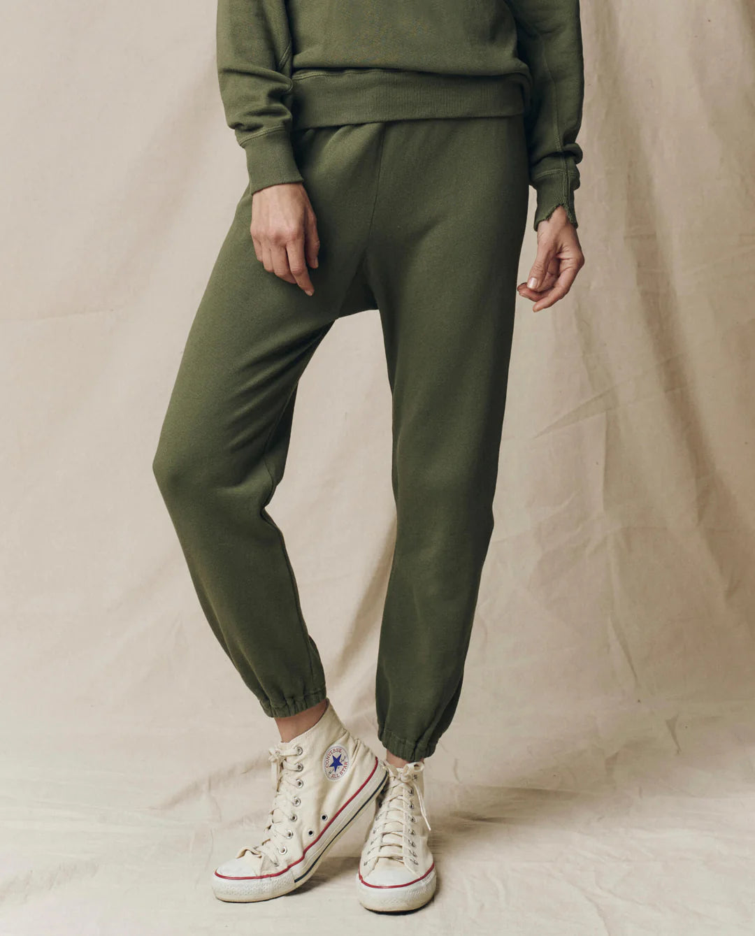 A person wears The Great Inc. Stadium Sweatpant in olive green with a matching sweatshirt and white high-top Converse, standing on a neutral fabric backdrop. Their face isn’t visible, evoking vintage athletic wear style.