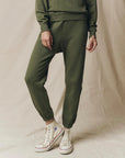 A person wears The Great Inc. Stadium Sweatpant in olive green with a matching sweatshirt and white high-top Converse, standing on a neutral fabric backdrop. Their face isn’t visible, evoking vintage athletic wear style.