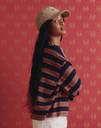 A woman with long black hair, wearing a beige cap and The Field Tee by The Great Inc., featuring navy blue, red, and white stripes, stands before a red patterned backdrop with her arms folded and head turned to the side.