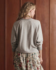 A person with long, wavy blonde hair faces a wooden wall, wearing The College Sweatshirt 2026 by The Great Inc. and a cream floral skirt with colorful botanical patterns—showcasing effortless handcrafted LA style.