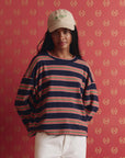 A person wearing a beige cap, The Great Inc. The Field Tee in navy blue stripes, and white pants stands against a red patterned background, looking at the camera.