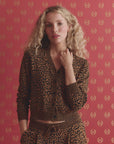 A woman with long curly blonde hair wears The Great Inc.'s The Shrunken Vneck Cardigan and matching pants, standing against a red patterned background with gold motifs. She faces forward, one hand touching her collar.