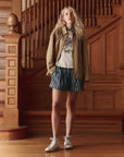 A person with long curly blonde hair stands indoors by a wooden staircase, wearing The Great Inc. The Folsom Jacket, a graphic T-shirt, blue-and-white striped shorts, and high-top sneakers.
