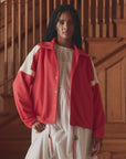 A person with long dark hair stands indoors by a wooden staircase, wearing The Trainer Jacket by The Great Inc. with white stripes over a white dress.