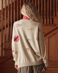 A person with long, wavy blonde hair stands on a wooden staircase, wearing The Great Inc.’s Embroidered Oversize Crew—an embroidered cotton-blend pullover with sports-themed patches, stripes, heart designs—and striped shorts.