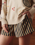 A person stands indoors with one hand in their pocket, wearing The Great Inc.'s The Boxer Short—tan, pull-on style with vertical black stripes—and a cream-colored sweater with embroidered patches.