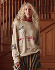 A young woman with curly blonde hair stands indoors by a wooden staircase, wearing The Great Inc.'s The Embroidered Oversize Crew in a soft cotton blend paired with black-and-tan striped shorts. She looks at the camera with a neutral expression.