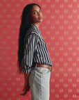 A woman with long dark hair stands sideways against red patterned wallpaper, wearing The Daily Button Up by The Great Inc.—an oversized black-and-white striped shirt—and light blue jeans, gazing at the camera with a relaxed expression.