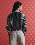 A person with long dark hair, seen from behind, wears The Daily Button Up by The Great Inc., featuring black-and-white stripes, paired with light blue jeans, standing before red wallpaper with a gold pattern.