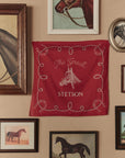 The Stetson Western Bandana by The Great Inc., featuring "The Great Stetson" and a horse head design, is displayed on a beige wall among framed horse paintings, adding vintage charm to any handkerchief-loving space.