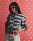 A woman with long braided hair wears The Great Inc.'s The Daily Button Up in black-and-white stripes and light blue jeans, standing against pink patterned wallpaper with a relaxed expression.