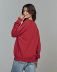 A woman with long brown hair stands sideways against a plain gray background, smiling slightly while wearing The Great Inc.’s oversized red College Sweatshirt S25 and light blue jeans.