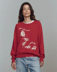 A woman wears The Great Inc.'s The College Sweatshirt S25, an oversized vintage top with a white "Merry Christmas" Santa and reindeer graphic, paired with light blue jeans, standing against a plain gray background.