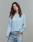A woman with long brown hair wears The College Sweatshirt S25 by The Great Inc. in light blue with a vintage "Happy Hanukkah" design. She smiles slightly, standing against a plain gray background with one hand in her jean pocket.