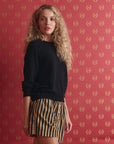 A woman with long curly blonde hair wears a black sweater and The Great Inc. The Boxer Short in beige with black vertical stripes, standing in front of red patterned wallpaper.