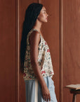 A woman with long braided hair stands in profile against a wooden wall, wearing The Victorian Pleat Tank by The Great Inc. in vintage rose print with light blue pants. An open book rests on the table beside her.
