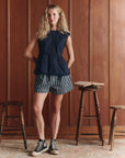 A woman with long curly blonde hair stands indoors by wood-paneled walls, wearing a sleeveless navy top, The Boxer Short by The Great Inc. in blue and white stripes, and black high-top sneakers. Two stools and an open book are nearby.