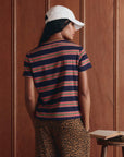 Wearing The Little Tee by The Great Inc. in navy blue stripes, paired with leopard print pants and a white cap, a person stands with their back to the camera before a wooden wall. An open book rests on a table nearby.