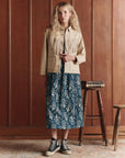 A woman with long, wavy blonde hair stands indoors against wood paneling, wearing The Great Inc. Leather Fringe Jacket over a blue floral midi dress and black high-top sneakers, beside a small stool stacked with books.